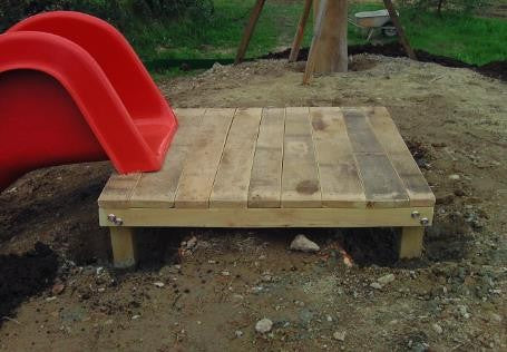 Wooden platform made from robinia wood in a garden setting with a red slide in the background.